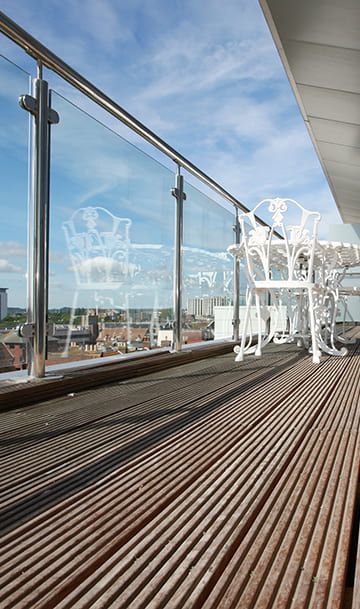Ein moderner Balkon mit polierten Holzböden, einem Glasgeländer und weißen Metallmöbeln vor einem klaren Himmel im Hintergrund.