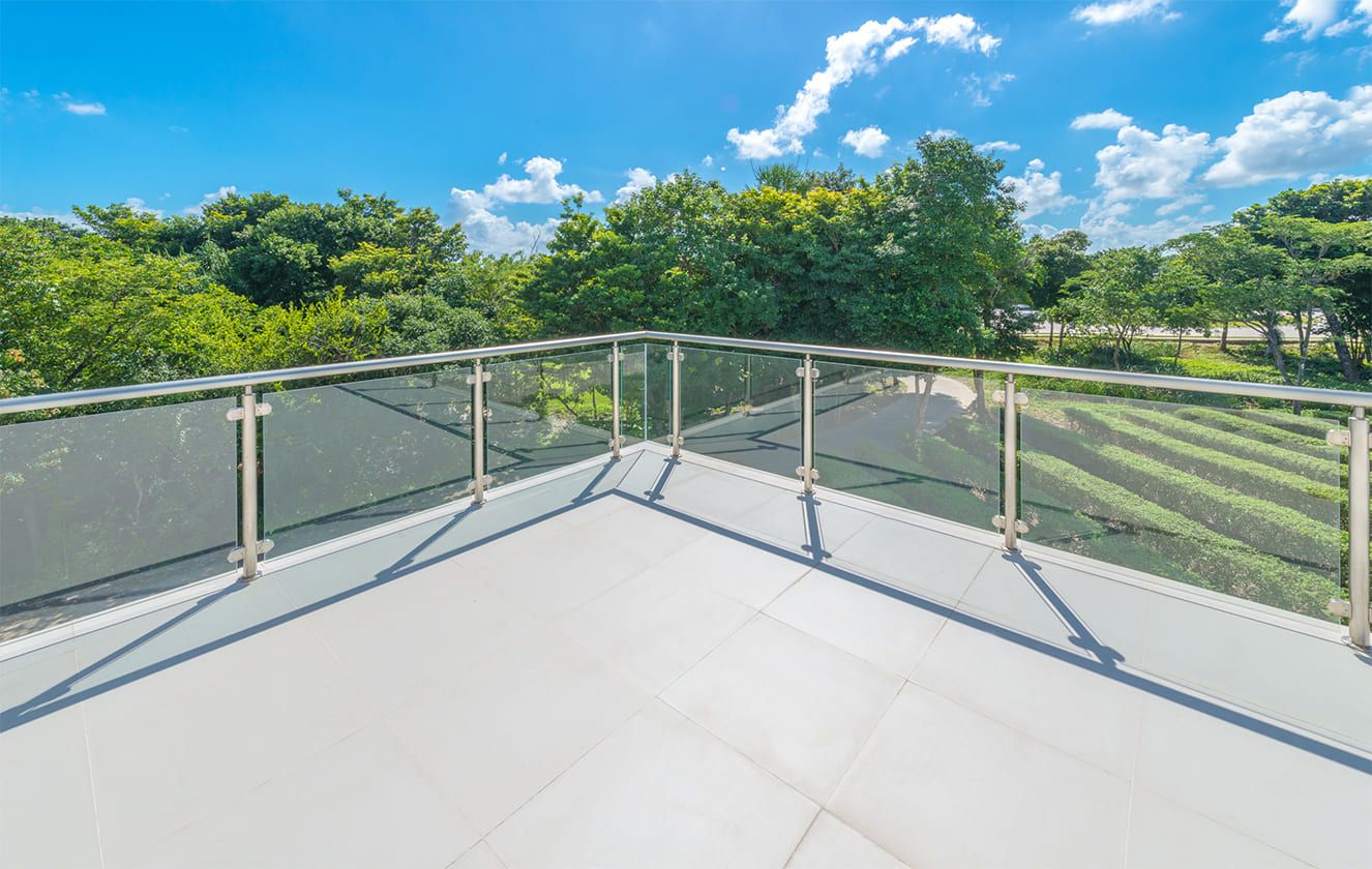 Sonniger Balkon mit Glasgeländer und Blick auf üppiges Grün und einen blauen, mit weißen Wolken übersäten Himmel. Ideal für Entspannung und Aussicht.