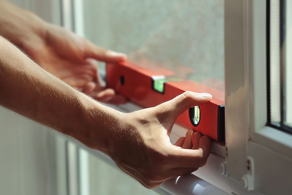Eine Person prüft mit einer roten Wasserwaage die Ausrichtung eines Fensterrahmens bei hellem Tageslicht.